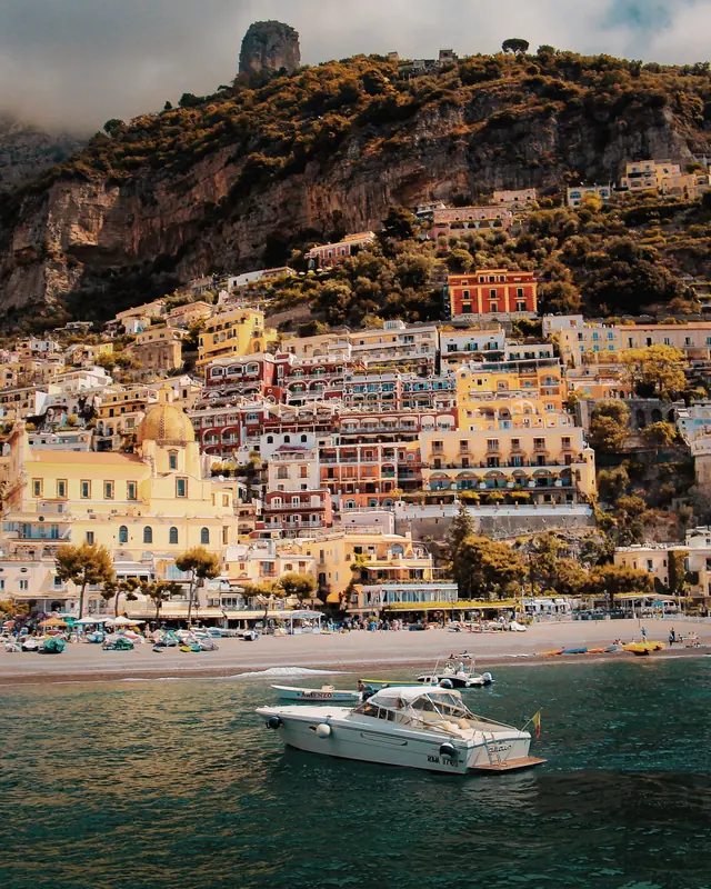 A white boat in front of a town stacked up a rocky cliff above a beach