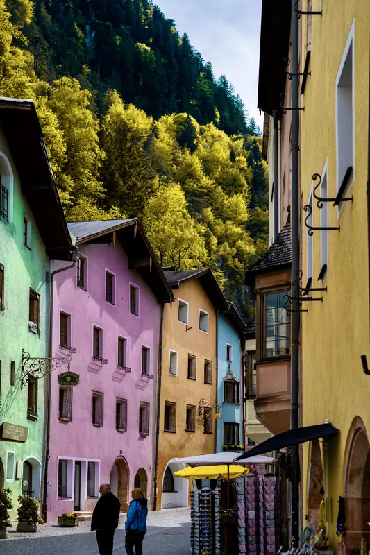 Two people on a cobbled street between pastel houses, with a bright green forest rising behind them